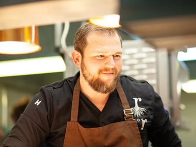das-excellence-gourmetfestival-10.jpg Mann im schwarzen Hemd und brauner Schürze in einer Küche.