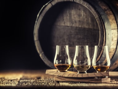 whisky-tasting-wenige-tropfen-offenbaren-unzaehlige-nuancen.jpg Drei Whiskygläser auf Tablett vor einem Fass.