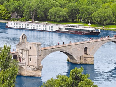 unterwegs-auf-dem-fluss.jpg Alte Steinbrücke über Fluss mit Kreuzfahrtschiff und Bäumen im Hintergrund.