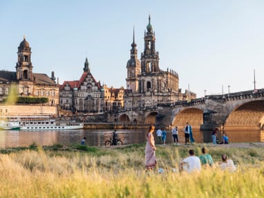 wahrzeichen-dresdens-die-augustusbruecke-und-die-frauenkirch.jpg Personen entspannen im Gras mit Blick auf historische Gebäude und eine Brücke am Flussufer.