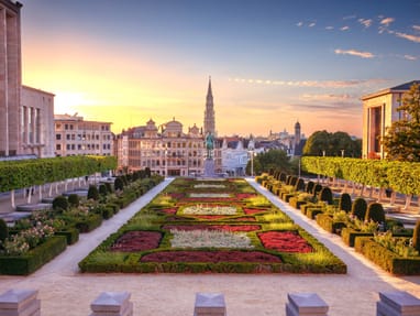 vom-kunstberg-mont-des-arts-oeffnet-sich-einer-der-schoenste.jpg Gartenanlage mit geometrischen Beeten und Stadtblick im Hintergrund bei Sonnenuntergang.