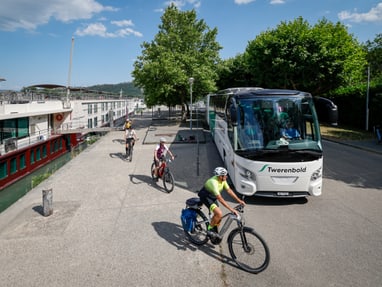 unterwegs-mit-schiff-bus-und-velo.jpg Menschen fahren mit Fahrrädern neben einem geparkten Reisebus und einem Flusskreuzfahrtschiff.