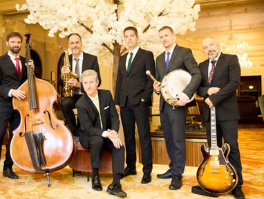 Sechs Männer in Anzügen mit Musikinstrumenten in einem eleganten Raum.