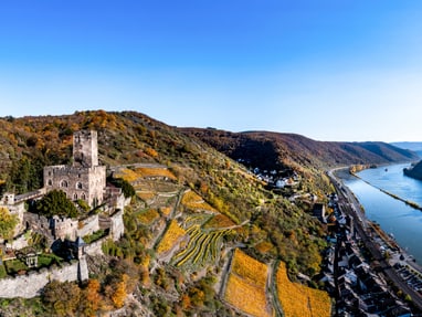 unesco-weltkulturerbe-oberes-mittelrheintal.jpg Luftaufnahme einer Burg mit Weinbergen und Fluss.