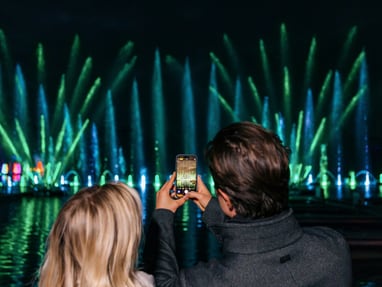 lichtergarten-illumina-im-schloss-laxenburg.jpg Menschen fotografieren Wasserfontänen-Show bei Nacht.