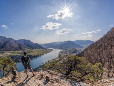 wachau.jpg Person auf einem Felsen mit Blick auf einen Fluss und Berge bei Sonnenschein.