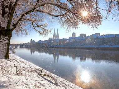 blick-auf-koeln-im-winter.jpg Verschneite Uferpromenade mit Bäumen und einer Stadt im Hintergrund, Sonne im Himmel.