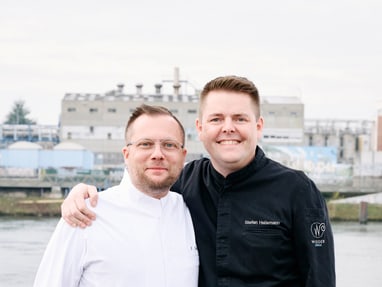 das-excellence-gourmetfestival-10.jpg Zwei Männer in Kochjacken vor industriellem Hintergrund.