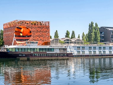 aufregend-modern-orange-cube-in-lyon.jpg Hausboot auf Fluss, moderne Gebäude im Hintergrund.