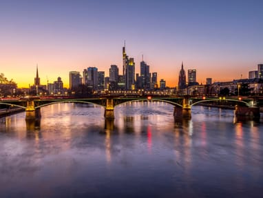 skyline-von-frankfurt-am-main.jpg Fluss vor Stadtansicht bei Sonnenuntergang mit beleuchteten Gebäuden und Brücken.