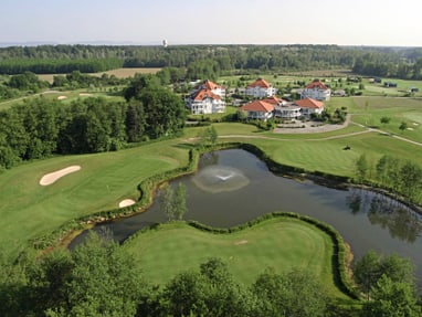Luftaufnahme eines Golfplatzes mit Teich und Gebäuden im Hintergrund.