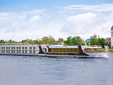 Grosses Flusskreuzfahrtschiff auf einem Fluss mit Bäumen und Turm im Hintergrund.