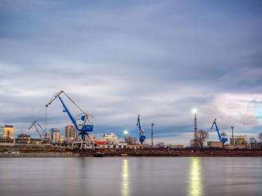 bratislava-aus-einer-anderen-perspektive-auf-der-faehrte-wah.jpg Flussufer mit Kränen und städtischer Skyline im Hintergrund unter bewölktem Himmel.