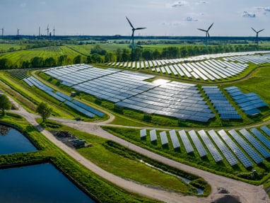 wind-und-solaranlagen-und-weitere-erneuerbare-energieliefera.jpg Luftaufnahme einer Solarfarm mit Windkraftanlagen und Teichen in einer grünen Landschaft.