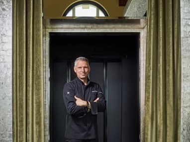 das-excellence-gourmetfestival-10.jpg Mann in schwarzer Kleidung steht mit verschränkten Armen vor Vorhang.