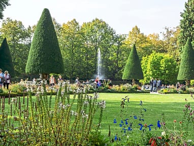 Menschen spazieren in einem gepflegten Garten mit Springbrunnen und Bäumen.