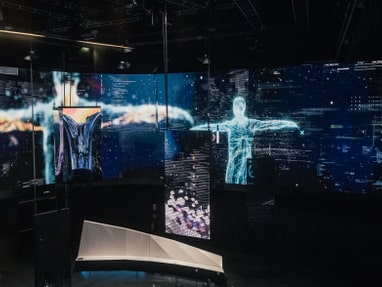 das-haus-der-digitalisierung-in-tulln-macht-digitale-themen.jpg Digitale Projektionen von Menschenfiguren und abstrakten Formen auf Bildschirmen in einem dunklen Raum.