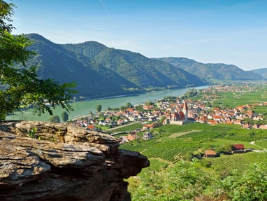 Blick auf ein Tal mit Fluss, Bergen und einer kleinen Stadt mit roten Dächern.