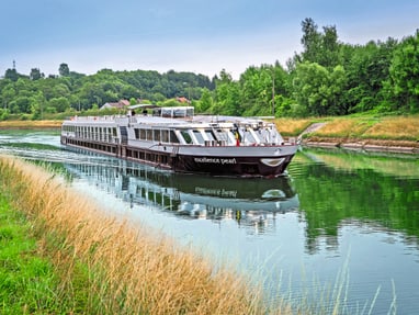 Das Flusskreuzfahrtschiff Excellence Pearl gleitet auf einem ruhigen Fluss durch eine grüne Uferland­schaft.