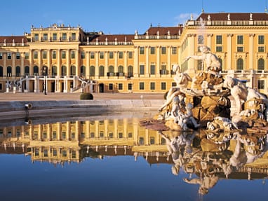 schloss-schoenbrunn-wien.jpg Historisches Gebäude mit gelber Fassade und einem Brunnen im Vordergrund, der sich im Wasser spiegelt.