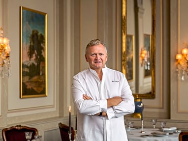 das-excellence-gourmetfestival-10.jpg Mann in weissem Hemd steht in einem eleganten Esszimmer mit Kerzen und Gemälden.
