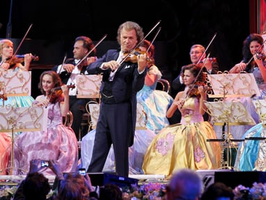 Geiger spielt mit Orchester auf Bühne in festlichen Kleidern.