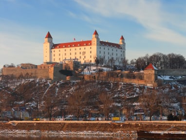aussicht-auf-die-burg-von-bratislava.jpg Ein Schloss auf einem Hügel mit Blick auf einen Fluss und Spiegelung im Wasser.