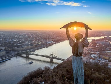 budapester-wahrzeichen-die-freiheitsstatue-auf-dem-gellertbe.jpg Monument über Budapest bei Sonnenuntergang mit Fluss und Brücke.