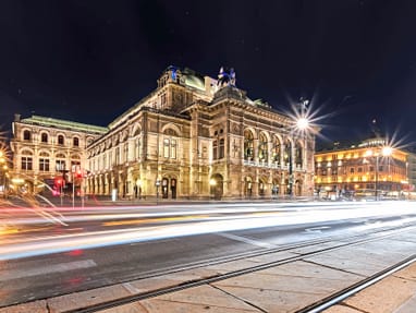 staatsoper-wien.jpg Beleuchtetes historisches Gebäude und bewegte Lichter von Autos bei Nacht.