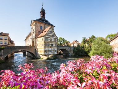 Historisches Gebäude auf einer Brücke über einem Fluss mit rosa Blumen im Vordergrund.