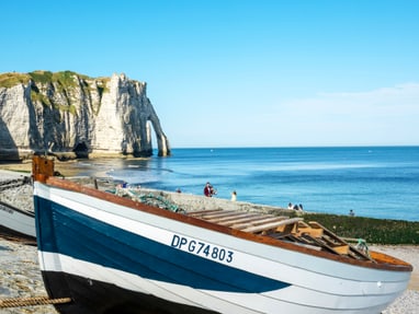 die-kreidefelsen-der-alabasterkueste-bei-etretat.jpg Strand mit Boot und Klippen im Hintergrund, klares blaues Meer.
