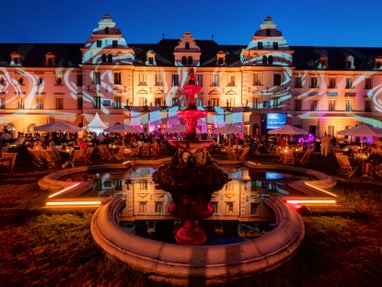 thurn-und-taxis-schlossfestspiele-regensburg.jpg Beleuchtetes Schloss bei Nacht mit Springbrunnen im Vordergrund.