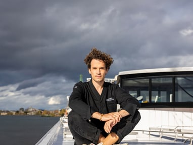 Person in schwarzer Kleidung sitzt auf einem Boot, mit bewölktem Himmel im Hintergrund.