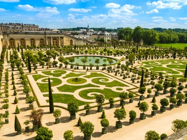 jardin-du-chateau-de-versailles.jpg Formaler Garten mit symmetrischen Hecken und Springbrunnen vor einem historischen Gebäude.