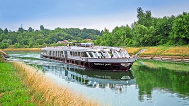 Flusskreuzfahrtschiff auf einem ruhigen Kanal mit grünen Ufern.