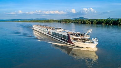 Flusskreuzfahrtschiff auf einem breiten Fluss mit bewaldetem Ufer.