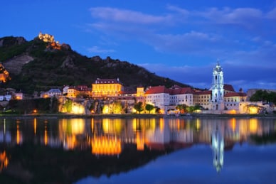 sommernaechte-an-der-donau-2.jpg Abendliche Flusslandschaft mit beleuchteten Gebäuden und Kirche.