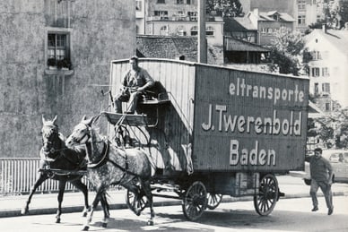Schwarz-Weiss-Foto eines Pferdegespanns mit geschlossenem Wagen der Firma J. Twerenbold auf einer Strasse in Baden, Schweiz.