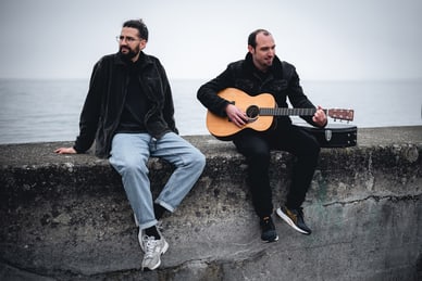 die-donau-alles-blueht-1.jpg Zwei Männer an der Küste, einer spielt Gitarre.
