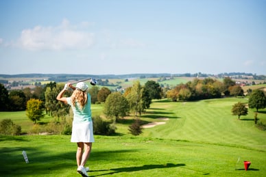 Frau in hellgrünem Shirt und weissem Rock führt einen Golfschlag auf einem weitläufigen Golfplatz aus.