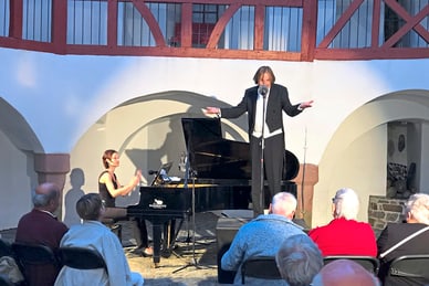 burkhard-von-puttkamer-und-seine-zwischenakt-kuenstlergruppe.jpg Musiker im Frack singt neben Pianistin auf Innenhofbühne vor Publikum.