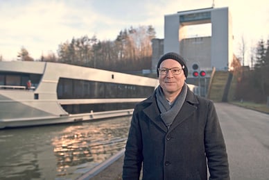grand-tour-basel-wien-1.jpg Mann steht vor einem modernen Flusskreuzfahrtschiff und einer Schleusenanlage.