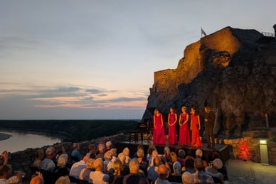 excellence-exklusiv-aus-purer-liebe-zur-musik-3.jpg Gruppe von Menschen sitzend, zuschauend, während einige Personen in roten Kleidern auf einer Bühne vor einer Burg performen.