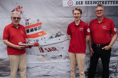 die-seenotretter.jpg Drei Personen in roten Hemden stehen vor einem Bild eines Rettungsboots.