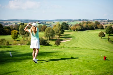 Frau in hellgrünem Shirt und weissem Rock führt einen Golfschlag auf einem weitläufigen Golfplatz aus.