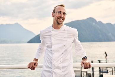 Mann in weissem Kochkittel steht draussen vor einem See mit Bergen im Hintergrund.