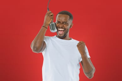 charles-nguela.jpg Person in weissem T-Shirt macht einen Luftspagat vor rotem Hintergrund und hält ein Mikrofon.