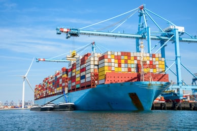 der-europoort-in-rotterdam-ist-europas-wichtiger-petrochemie.jpg Grosses Containerschiff im Hafen mit blauen Kränen und Windkraftanlage im Hintergrund.