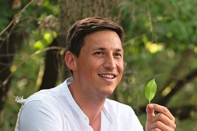 Kevin Nobs im weissen Hemd, lächelnd, hält ein grünes Blatt in der Hand, vor grünem Laubhintergrund.