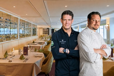 das-excellence-gourmetfestival-5.png Zwei Männer in Kochjacken stehen in einem eleganten Restaurant mit gedeckten Tischen.